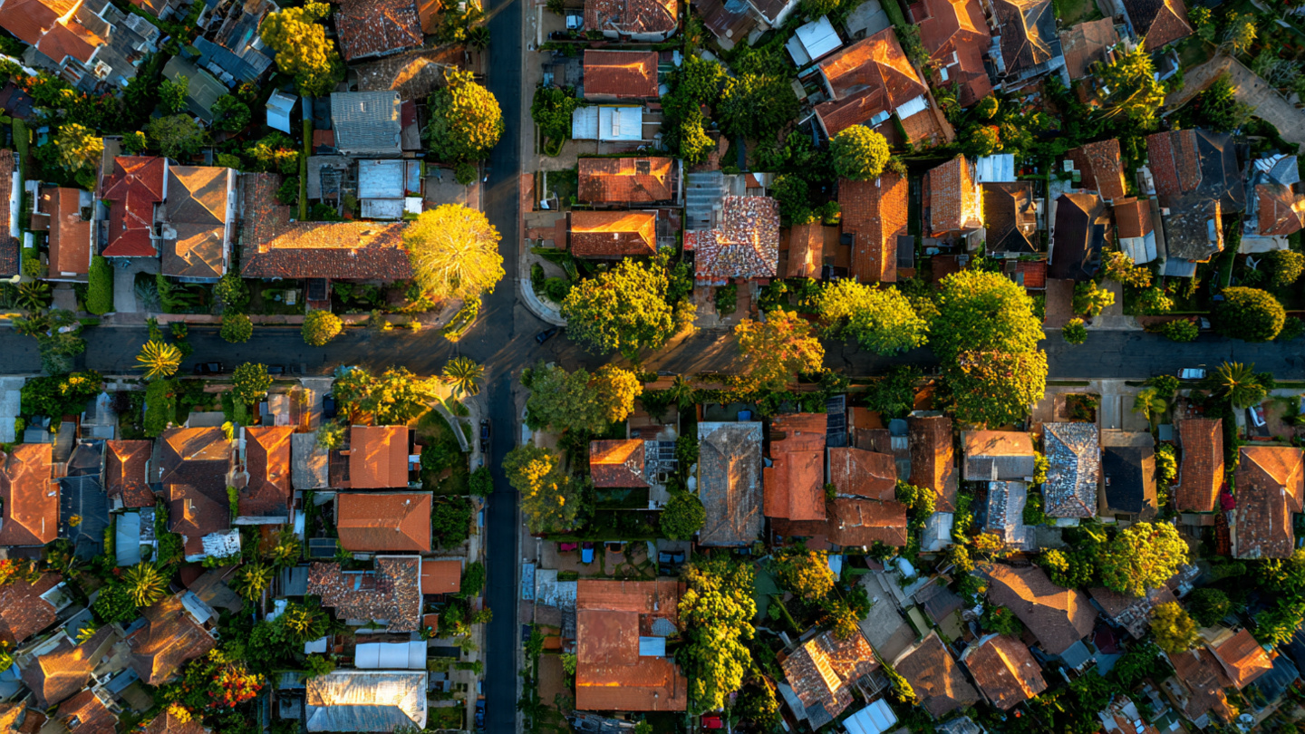 Aerial view of suburban residential rooftops in varying conditions, some with visible wear and patching, shot from directly above as if from a satellite or drone, warm late-afternoon light casting long shadows