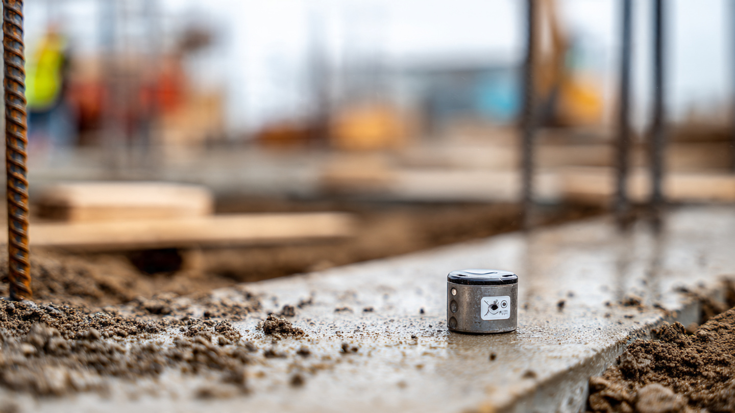 A small wireless sensor zip-tied to rebar inside a residential foundation form, fresh concrete being poured around it from a chute, morning light on a suburban job site