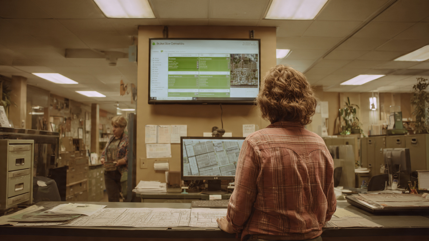 A homeowner standing at a city planning counter reviewing blueprints while a computer screen behind the clerk shows an AI zoning compliance interface with green checkmarks, warm overhead fluorescent lighting